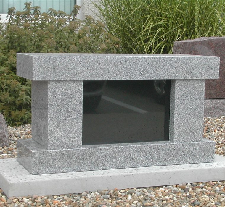 Grey granite cremation memorial with a polished black insert panel, set on gravel near garden shrubs.